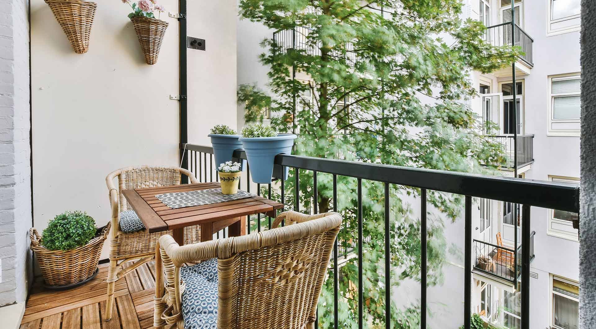 Balcón con sillas de mimbre, mesa pequeña, plantas en macetas y barandilla negra con vistas a la vegetación.